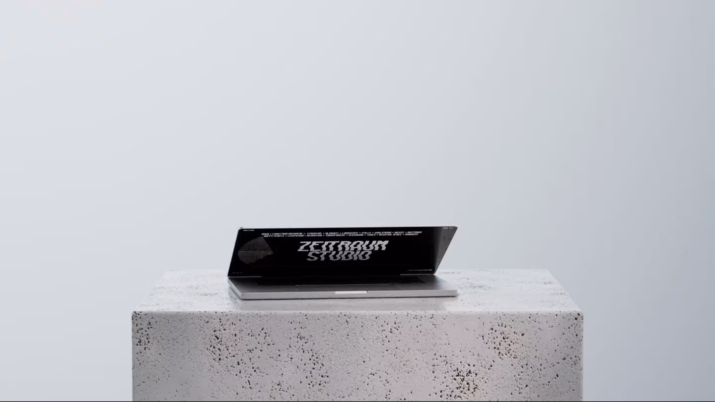 Partially open laptop displaying text "ZENTRAUM STUDIO" on its screen, placed on a textured concrete pedestal against a plain background.
